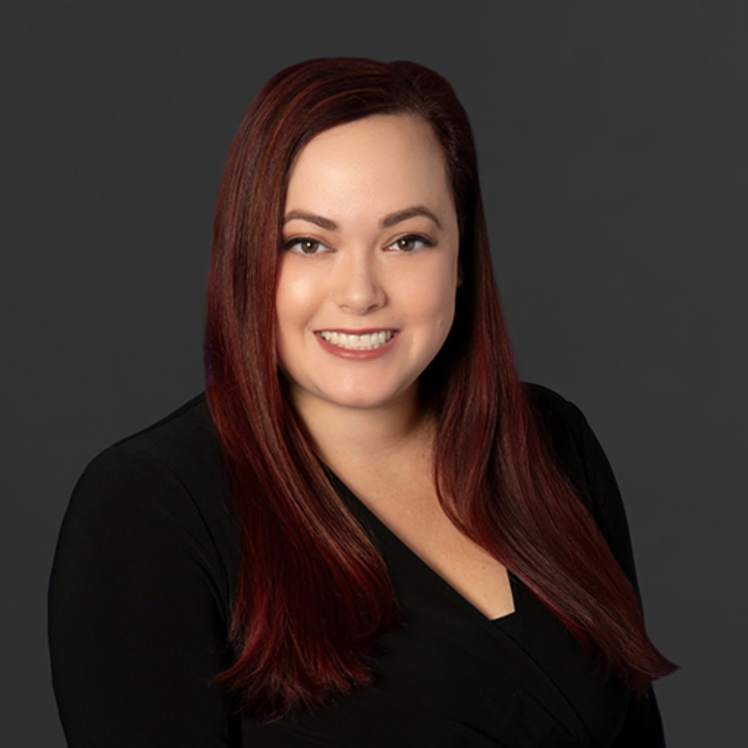 Headshot of a woman with long reddish-brown hair, smiling, wearing a black top, against a dark gray background.