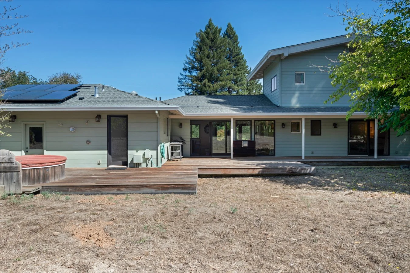 Rear view of a house with a wooden deck, solar panels on the roof, green exterior siding, large sliding glass doors, and outdoor furniture. There's a tree on the right side and tall trees in the background.