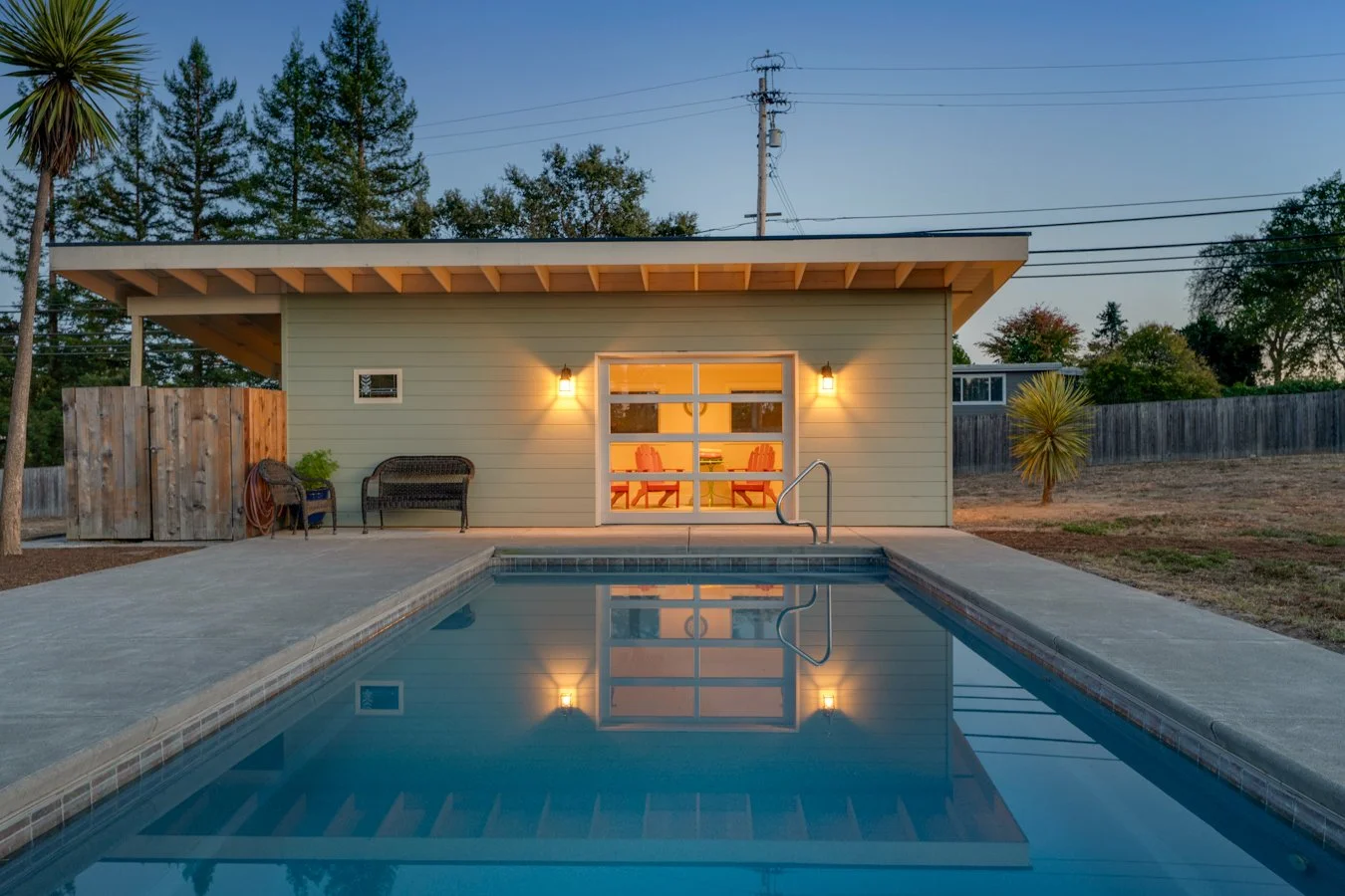 Backyard with a swimming pool, outdoor seating, small shed, and house with large glass doors and warm interior lighting at dusk.