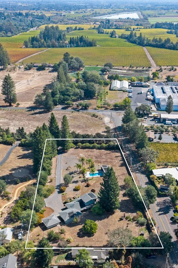 Aerial view of a residential property with a house, trees, a driveway, and a backyard containing a pool. The lot is outlined with a white border, with surrounding urban and rural landscapes, including fields, a commercial building, and a river in the