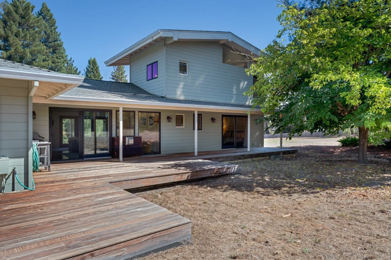 Exterior view of a two-story house with a wooden deck, sliding glass doors, a picnic table, and a lush green tree in the backyard, under a clear blue sky.
