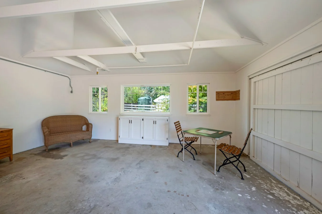 Interior of a garage with three small windows, a wicker loveseat, a small wooden dresser, a green table with two wooden chairs, and a white cabinet under the middle window. The garage floor is unfinished concrete.