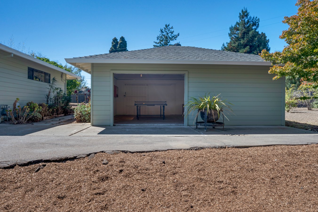 A white garage with an open door, a potted plant on a stand in front, and a foosball table inside. The garage is part of a house with a gray roof, surrounded by trees and bushes on a sunny day.