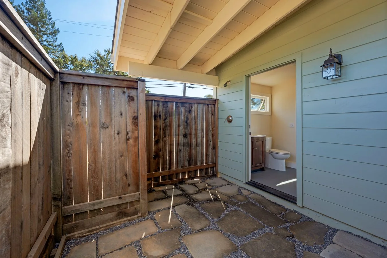 Small outdoor patio area with stone pavers, a wooden privacy fence, and a view of a bathroom with a toilet and a vanity through an open sliding door, under a covered roof.