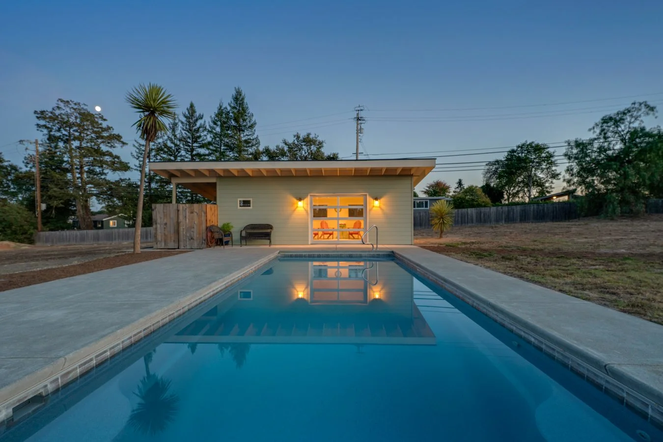 A backyard with a swimming pool, small house with outdoor seating, and trees under a dusk sky with the moon visible.