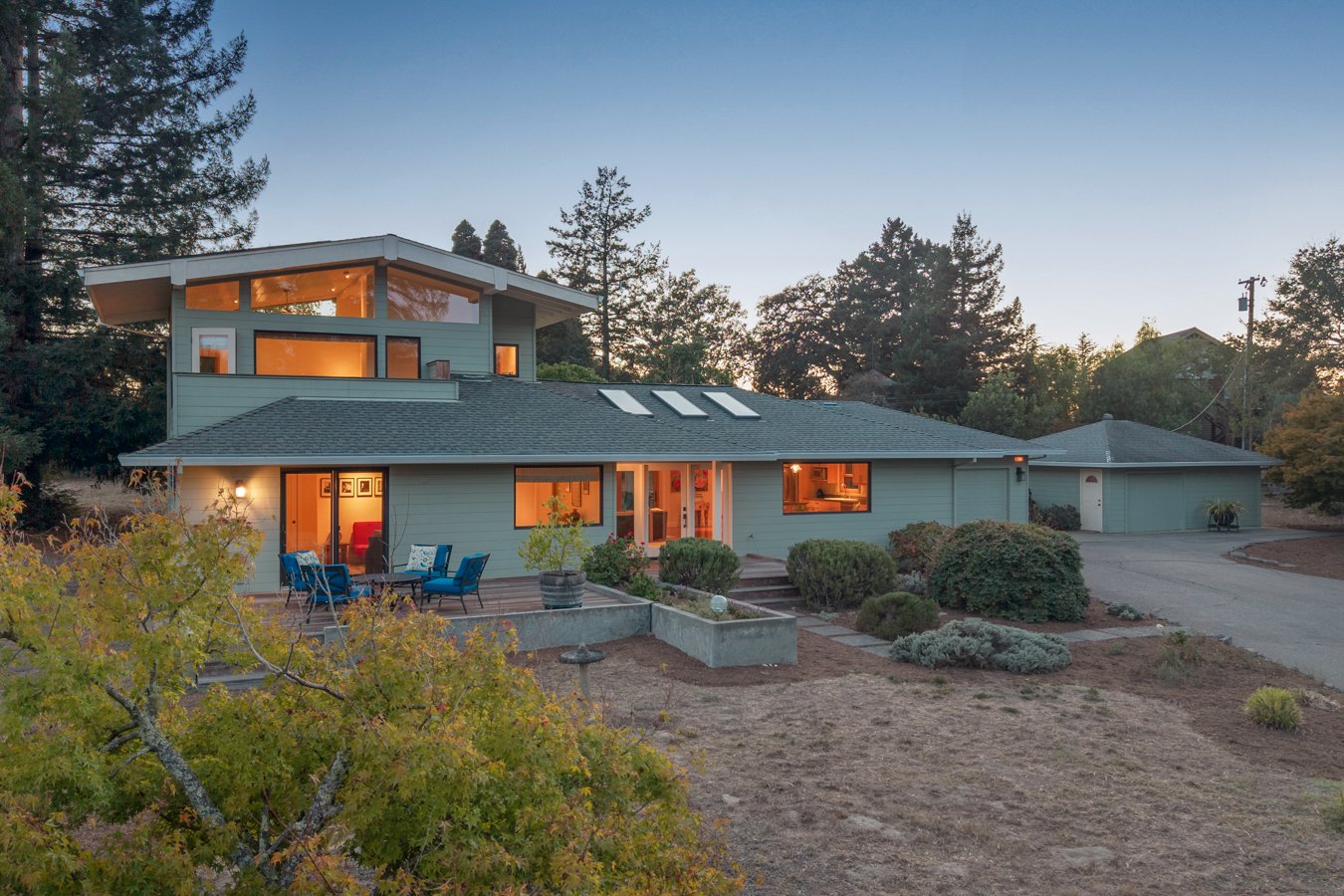 Modern two-story house with large windows and a patio with blue chairs, surrounded by trees and a landscaped yard, during sunset.