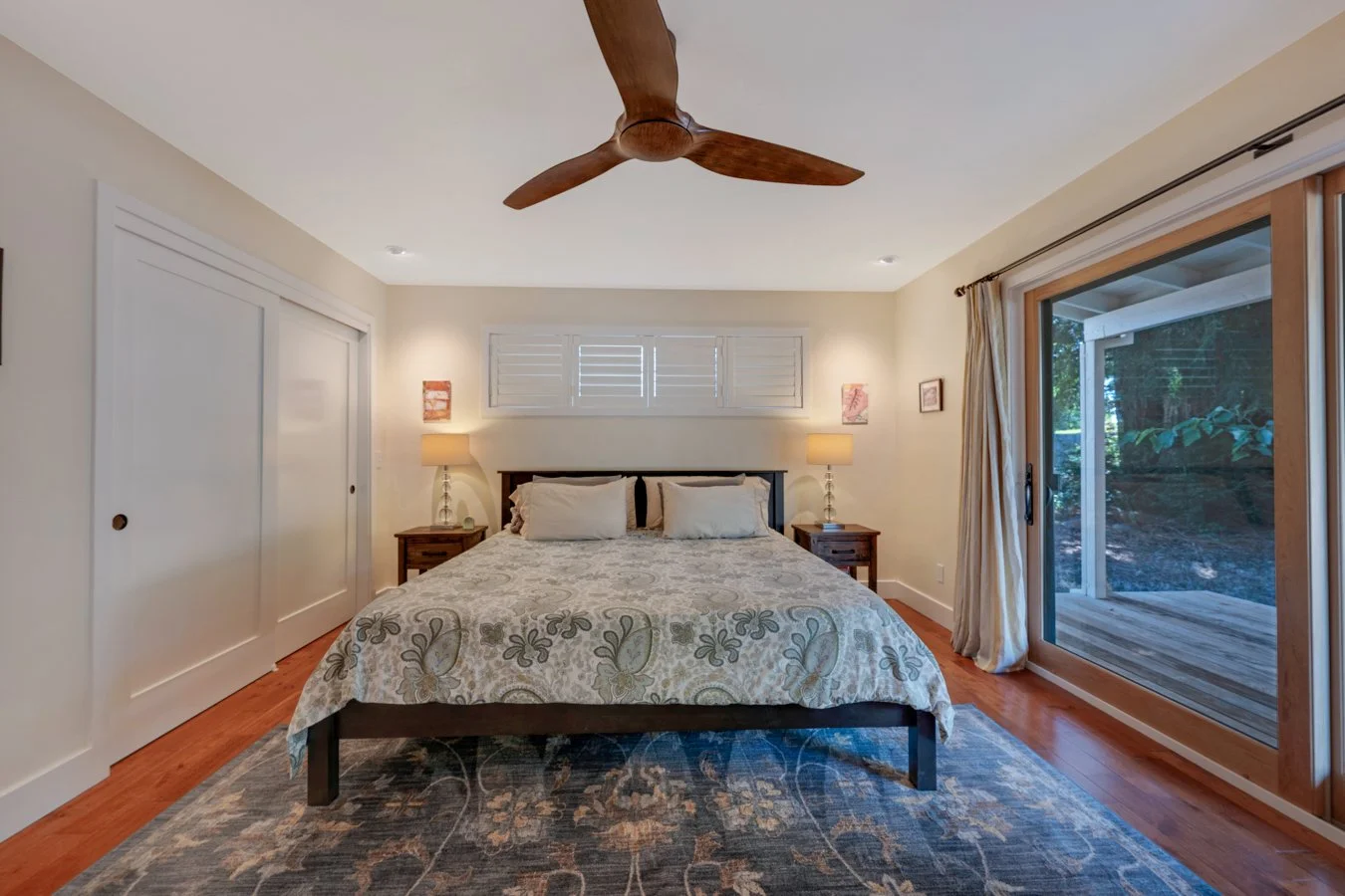 A bedroom with a bed, two bedside tables with lamps, a sliding glass door showing a wooden deck and trees outside, a ceiling fan, and a window with shutters.