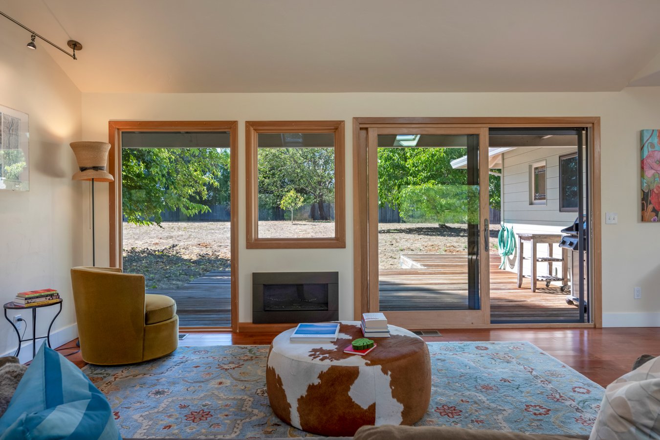 Living room with large glass sliding doors and windows, showing a backyard with trees and a deck outside. The room has a mustard yellow chair, a patterned area rug, a cowhide ottoman with books and a green coaster on top, and a small side table with 