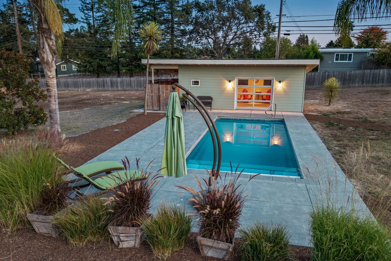 A backyard with a small pool, surrounded by plants, with a green lounge chair and umbrella on a patio. There is a small building with lights on, and trees and houses in the background.