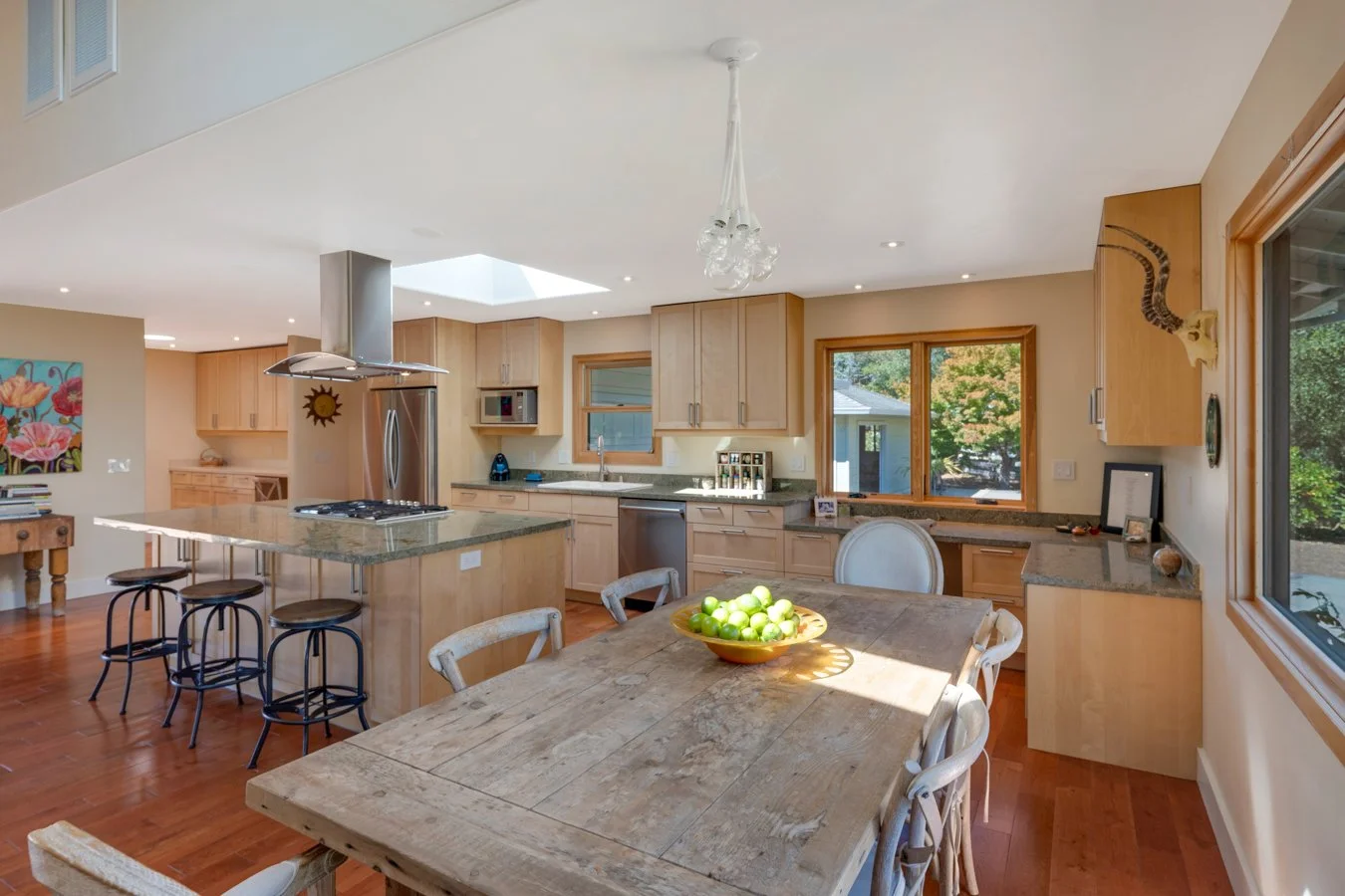 Bright kitchen with wooden cabinets, stainless steel appliances, a wooden dining table with a bowl of green apples, large windows, and a skylight, decorated with a mounted animal skull on the wall.