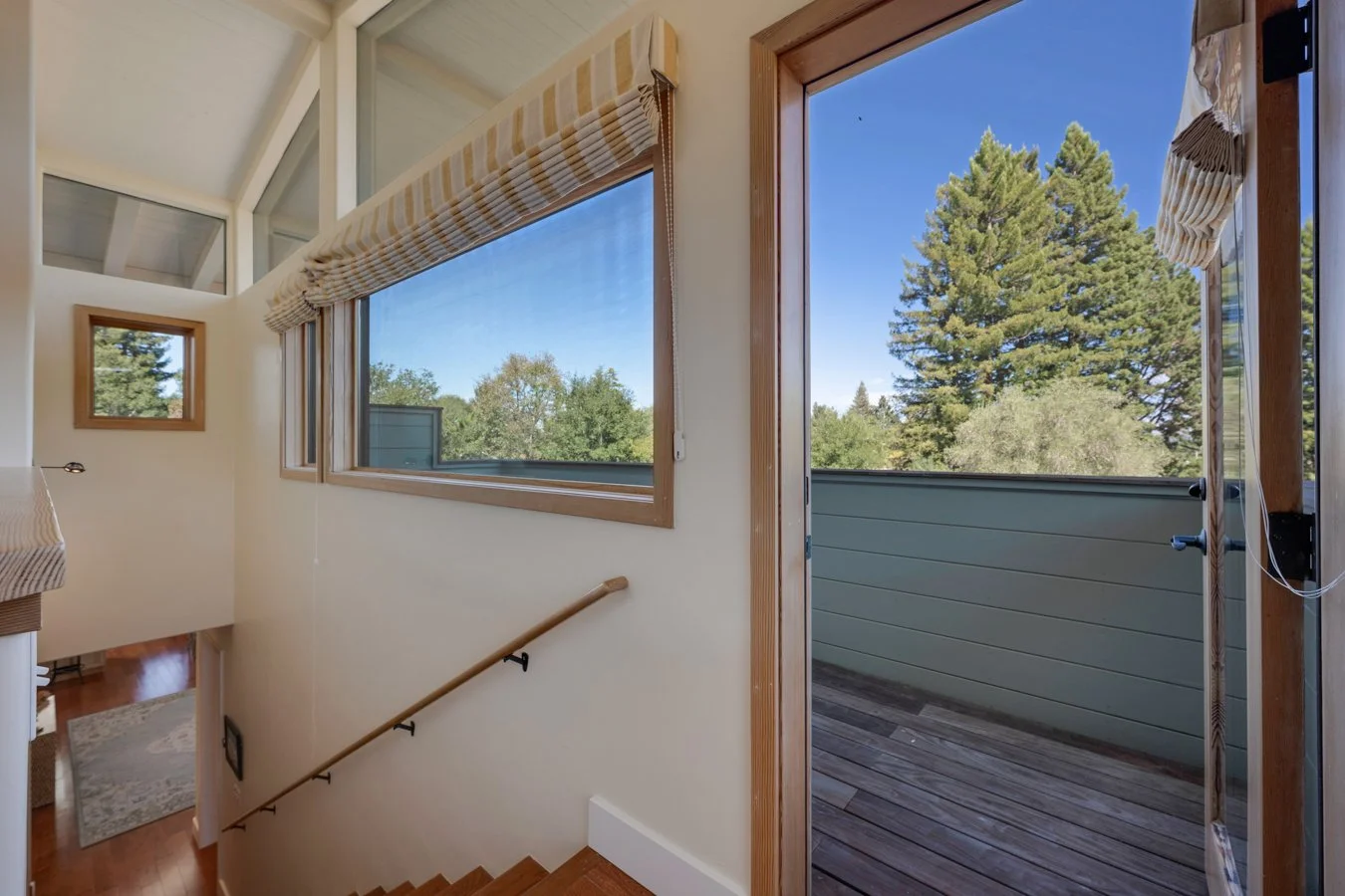 Interior view of a house showing a staircase leading downstairs, large windows with blinds, and a door opening to a balcony with a view of trees and a blue sky.