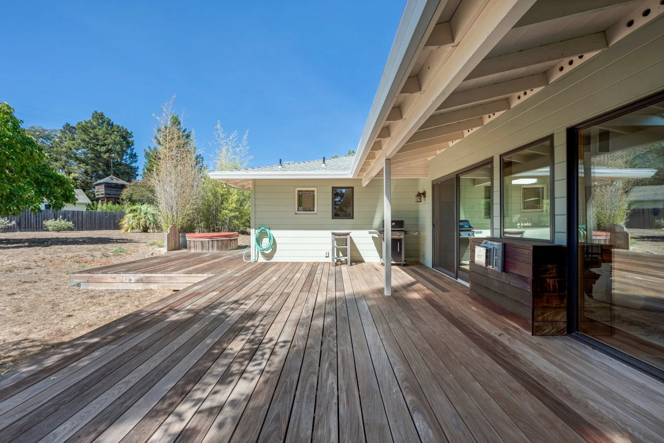 Wooden backyard deck with sliding glass doors, outdoor grill, and garden hose against house wall, with trees and a wooden fence in the yard and a clear blue sky overhead.