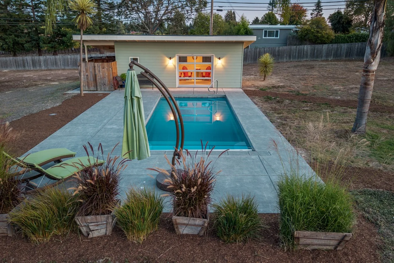 A backyard with a swimming pool, poolside lounge chair, and a small detached building with interior lighting visible through the window.