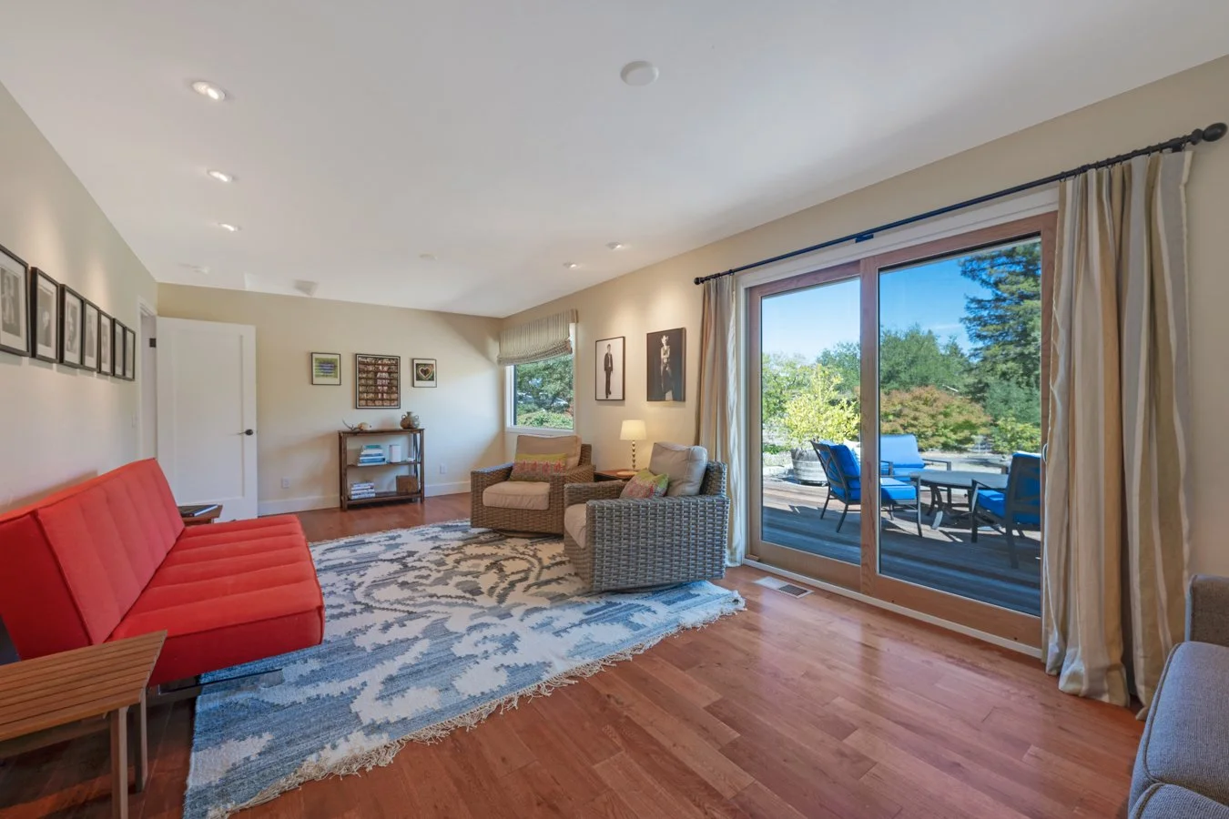 Living room with beige walls, hardwood floors, a red sofa, beige and patterned armchairs, a blue and white rug, and sliding glass doors leading to a patio with outdoor seating and trees in the background.