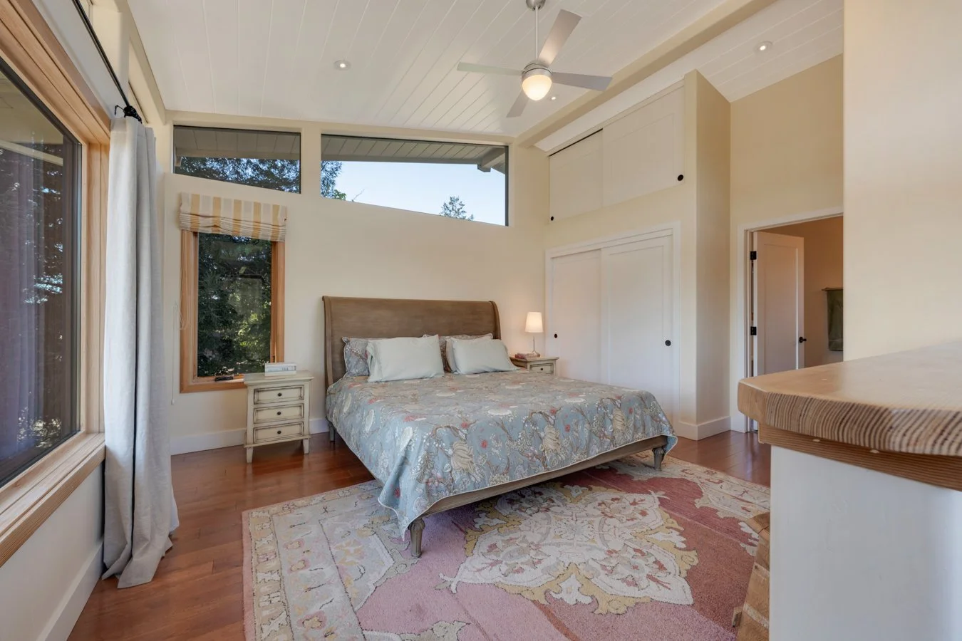 Beach style bedroom with a bed having a padded headboard, white pillows, and floral bedding. Large windows, ceiling fan, and a rug on wood flooring.