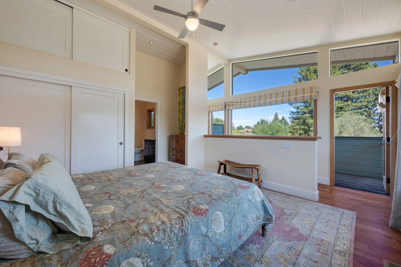  bedroom with large windows, a bed with floral bedding, a small bench, a ceiling fan, and a view of trees outside