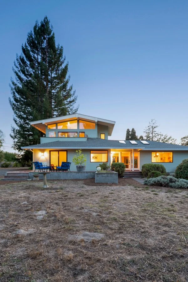 A modern two-story house with large windows, a sloped roof, and outdoor patio seating, surrounded by trees and a barren yard during twilight.