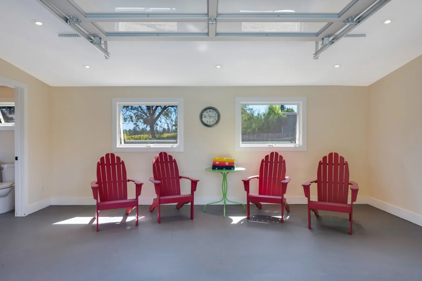 Bright garage converted into a sitting area with five red Adirondack chairs, a small green table with colorful stacked towels or blankets, two windows, and a wall clock.