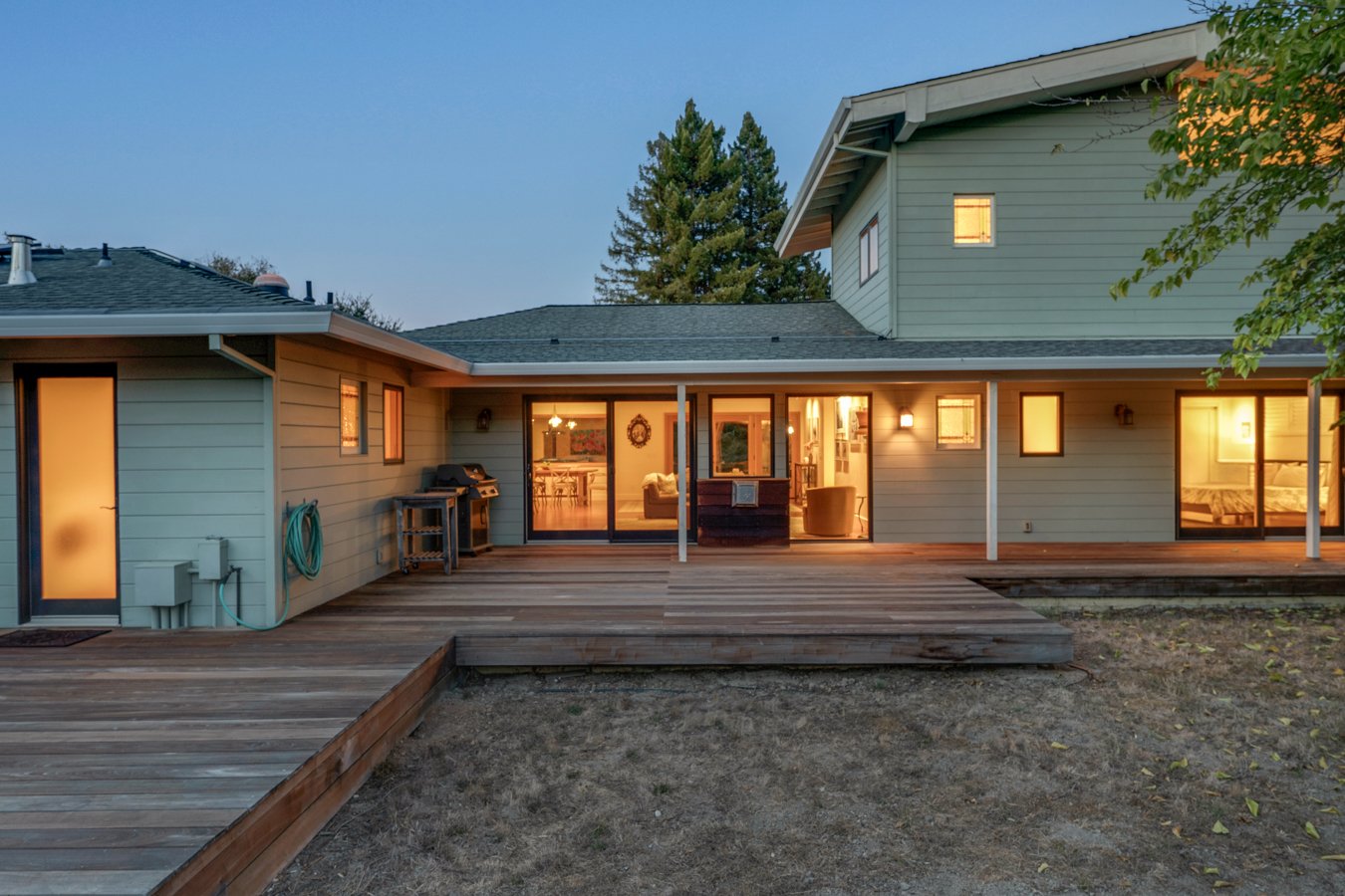 A two-story house with a wooden deck outside, illuminated from inside during dusk, with large glass sliding doors and windows, and trees in the background.