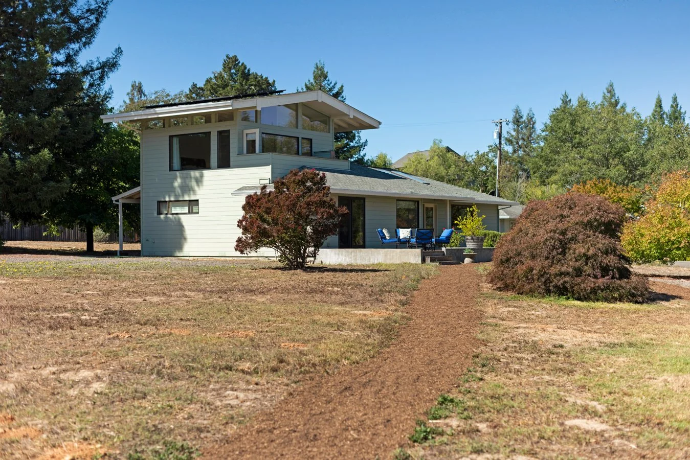 Modern two-story house with gray siding, large windows, a patio with blue chairs, and a dirt path leading to the porch, surrounded by trees and shrubs under a clear blue sky.