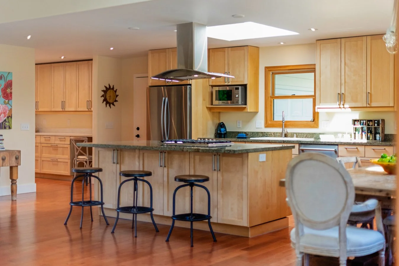 Bright kitchen with wooden cabinetry, stainless steel appliances, a central island with barstools, and a dining area with a white upholstered chair.