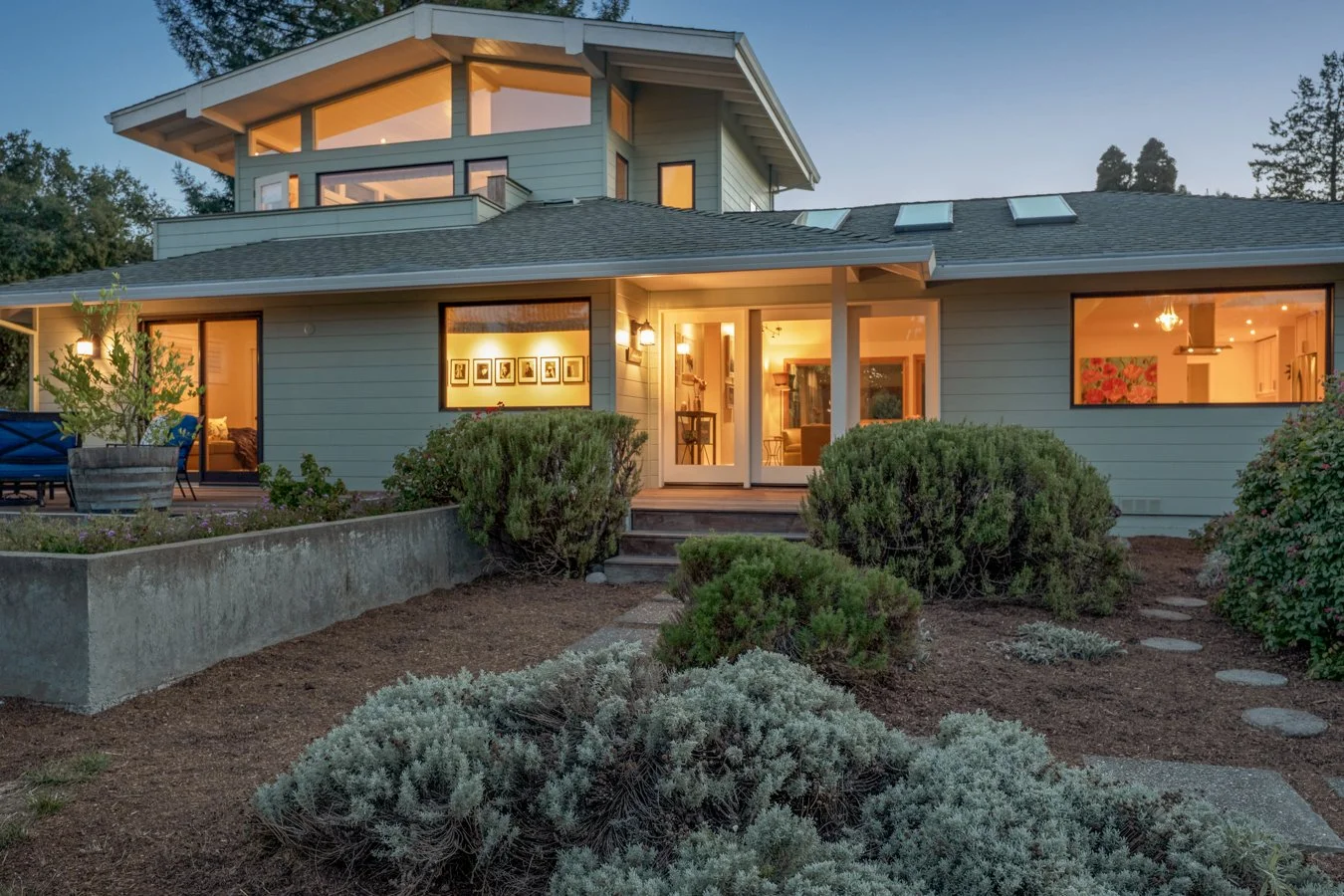 Modern house with large windows and a garden at dusk, warm interior lighting visible through the windows.
