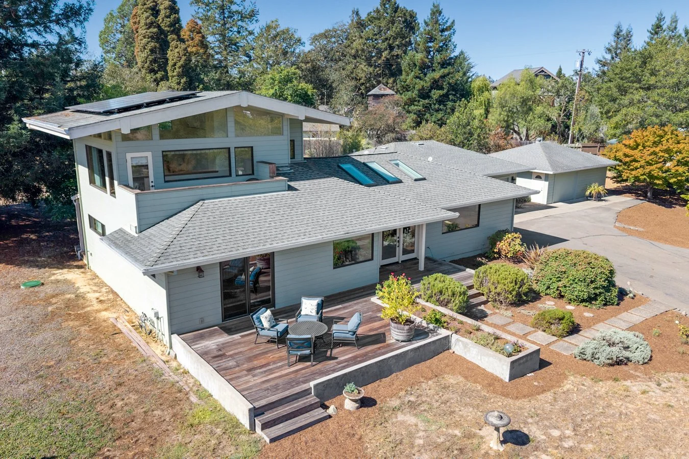 A modern two-story house with a wooden deck, outdoor seating, and solar panels on the roof, surrounded by trees and a yard with various plants.