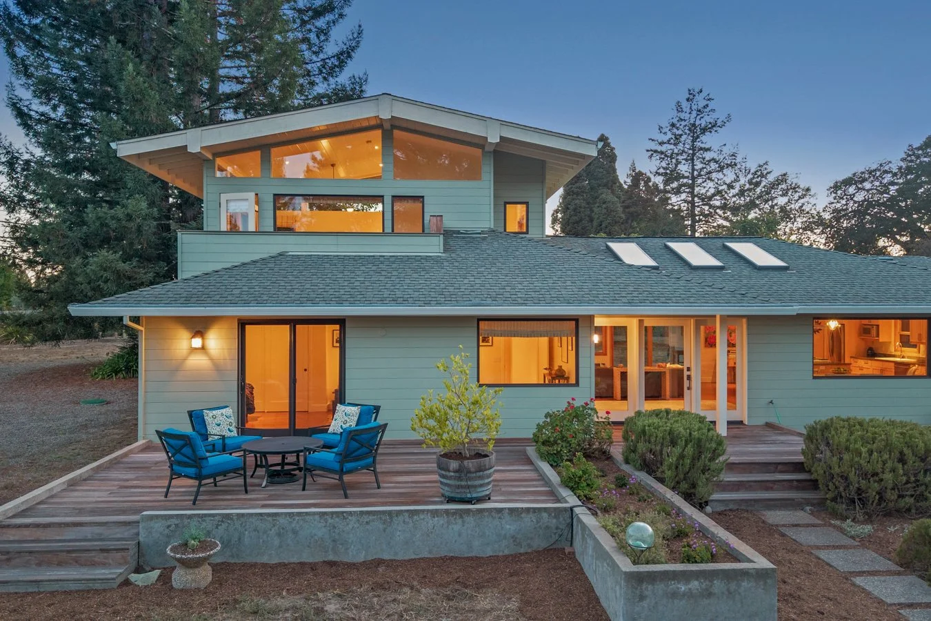 A modern two-story house with a wooden deck, outdoor seating, and large windows illuminated from inside, surrounded by trees at dusk.