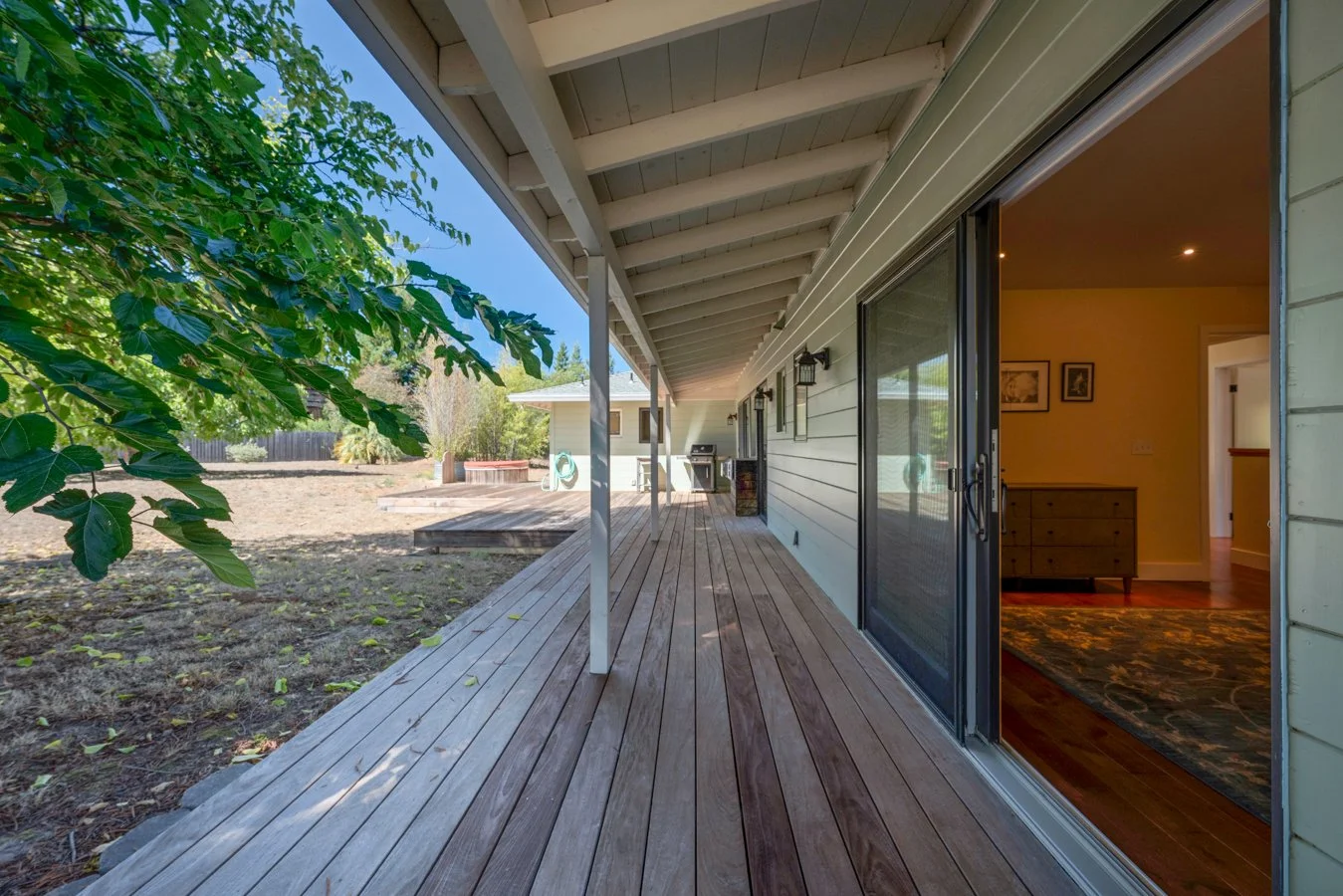 View of a wooden backyard deck with an open sliding glass door leading into a home, and a yard with a tree, a hot tub, and outdoor furniture in the background.
