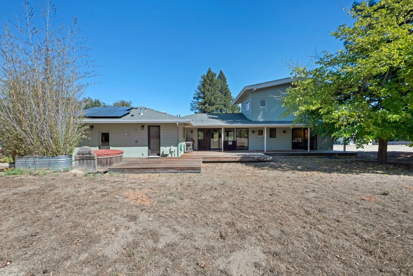 Back view of a house with a wooden deck, surrounded by a yard with trees, against a clear blue sky.