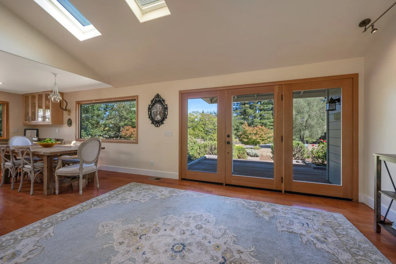 Living room with large glass sliding doors leading to an outdoor deck, wooden flooring, a patterned rug, dining area with table and chairs, and windows showing a garden outside.