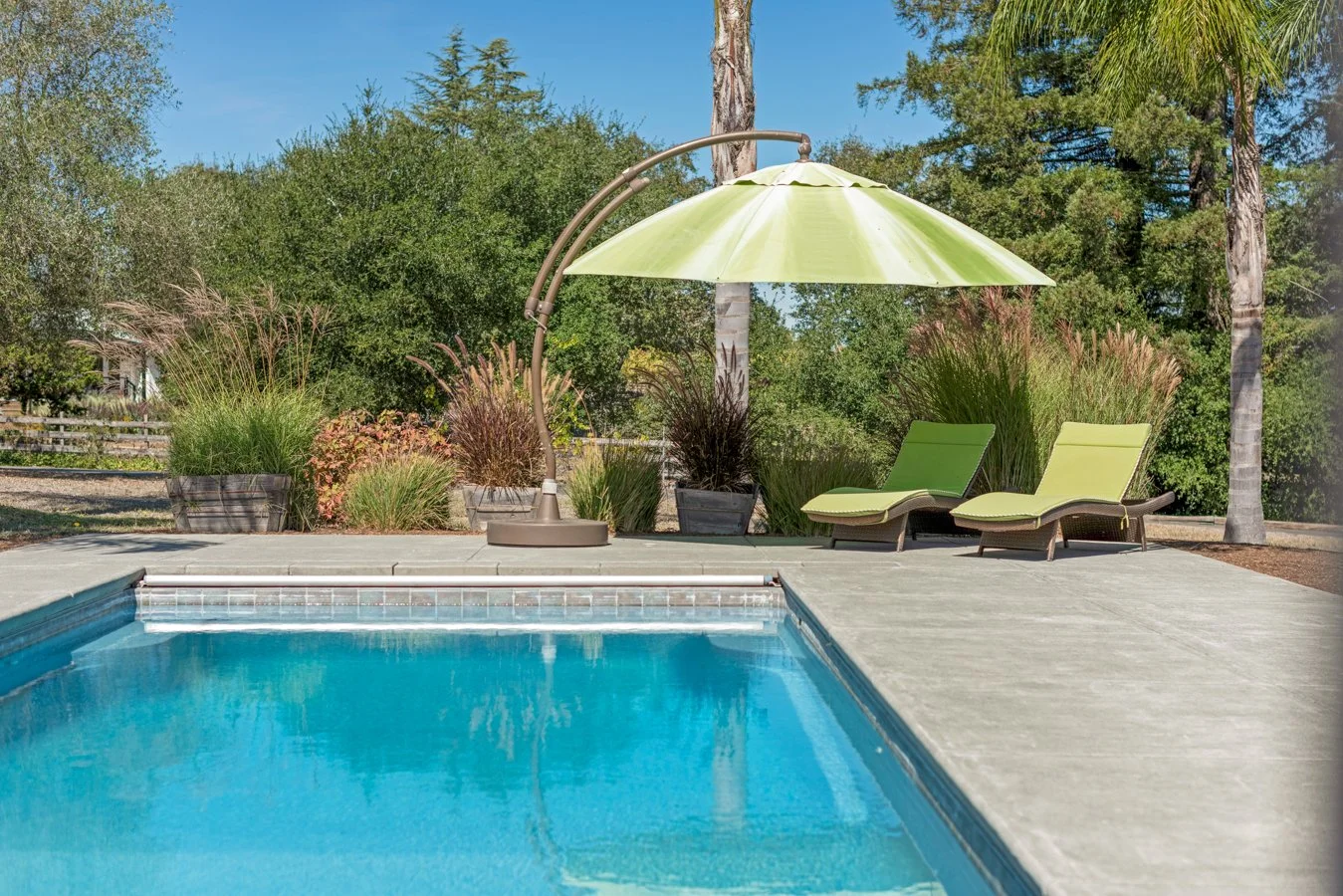 A backyard swimming pool with two green lounge chairs and a large green patio umbrella on a concrete deck, surrounded by trees and potted plants.