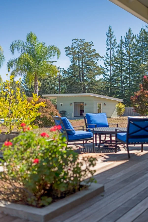 A backyard patio with blue cushioned chairs around a small table, a garden bed with pink flowers in the foreground, and a grass yard with a small beige house and tall trees in the background under a clear blue sky.