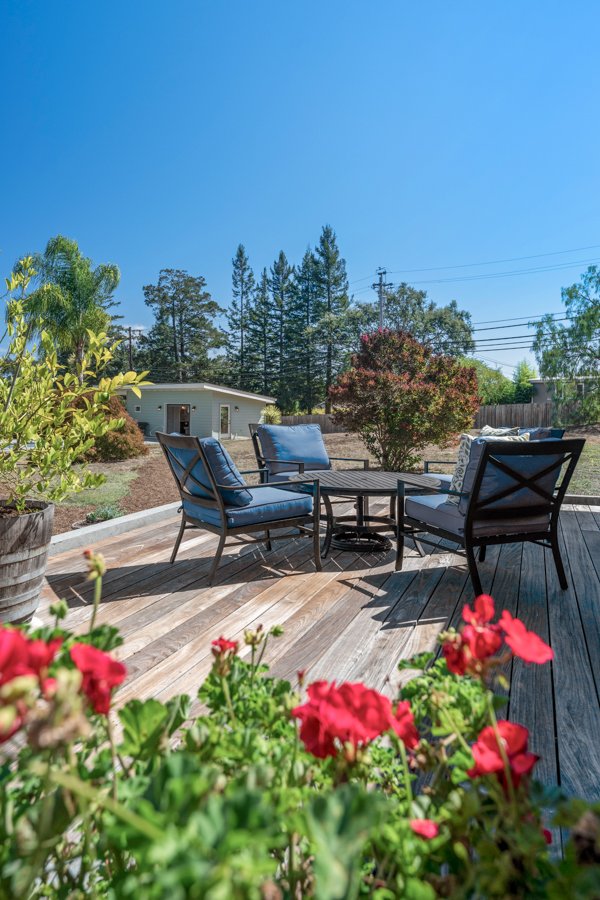 A backyard patio with a wooden deck, outdoor chairs with cushions, and flowering plants in the foreground under a clear blue sky.