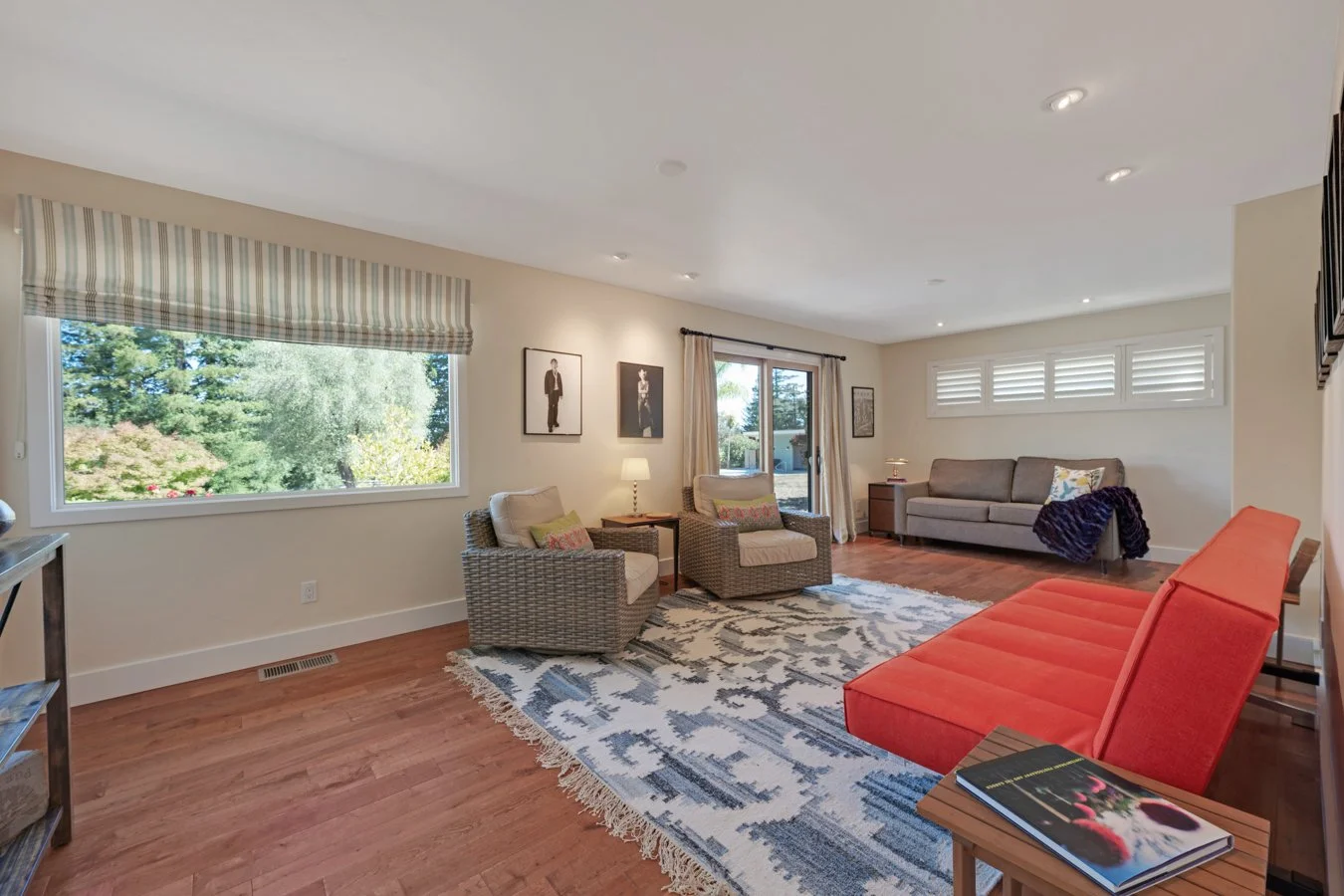 Living room with large window, neutral-colored walls, wooden flooring, various sofas including a bright orange one, a patterned rug, and framed artwork on the wall.