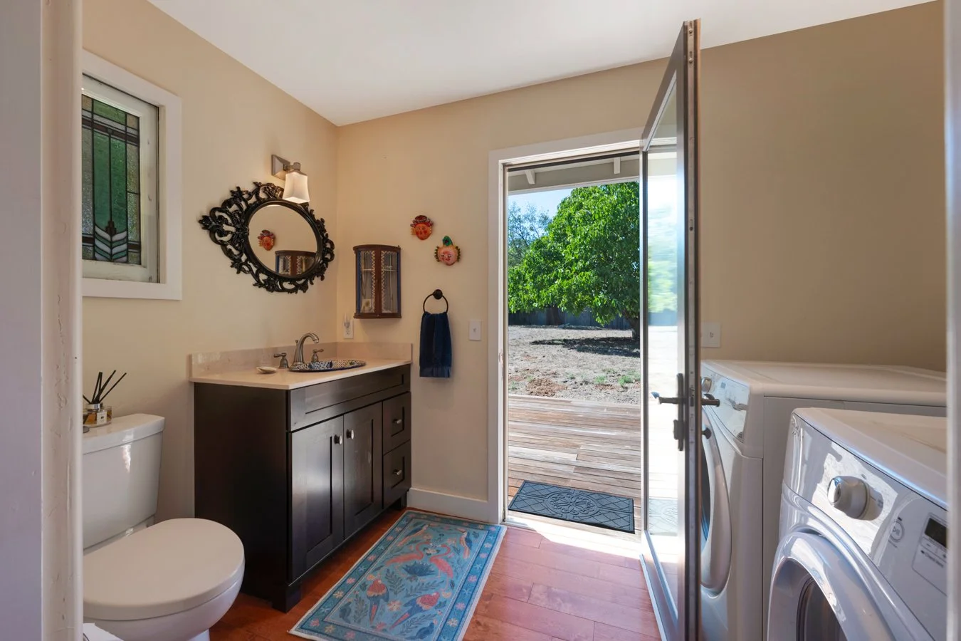 Laundry room with washer and dryer, a dark wooden cabinet with a sink, a decorative mirror, a small window, a wall-mounted cabinet, a blue towel, colorful wall decorations, a door leading outside to a wooden deck, and a blue rug with a flamingo desig