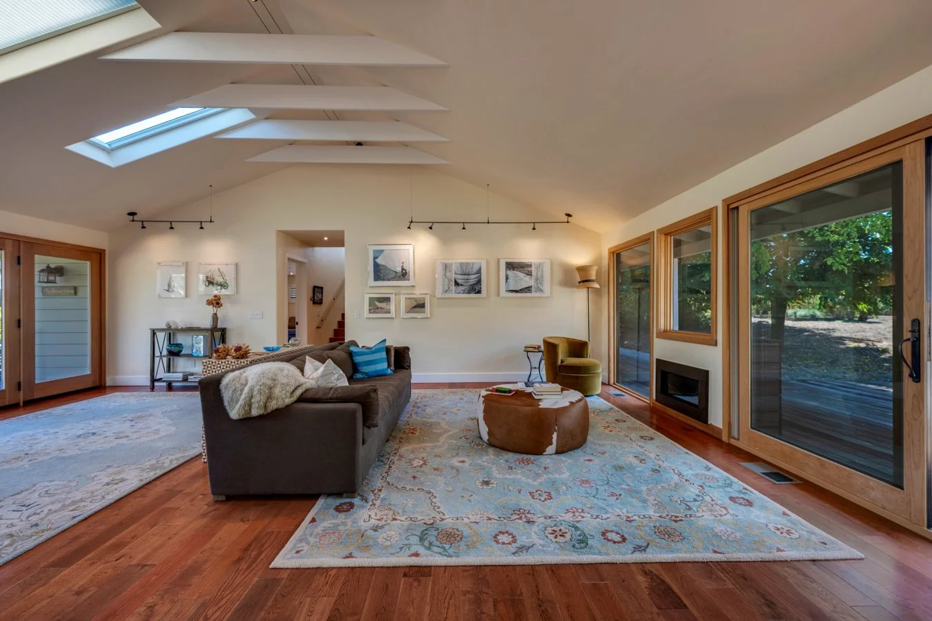 Spacious living room with hardwood floors, a large sofa with pillows and a throw blanket, a round ottoman, a green armchair, a rug, and large glass sliding doors leading outside. Artwork hangs on the wall, and there's a skylight in the ceiling.