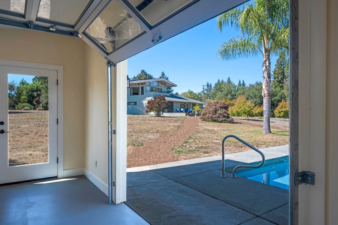 View from a room with open garage door showing a backyard with a swimming pool, a tree, and a house in the distance under a clear blue sky.