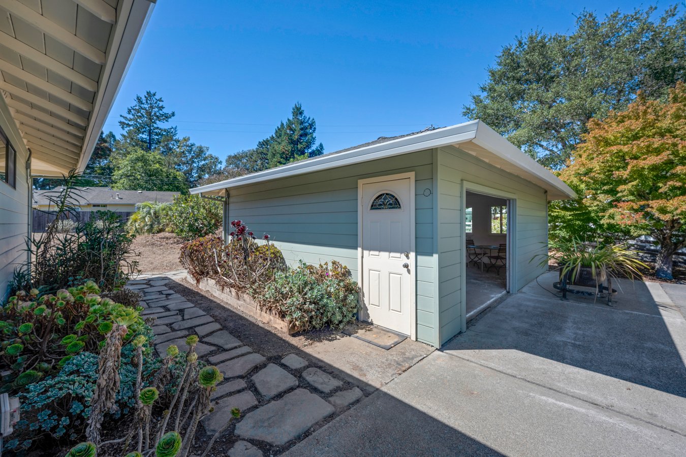 View of a small, light green shed with a white door and a window, situated in a backyard on a sunny day. Surrounding the shed are various plants and a concrete patio, with trees and a blue sky in the background.