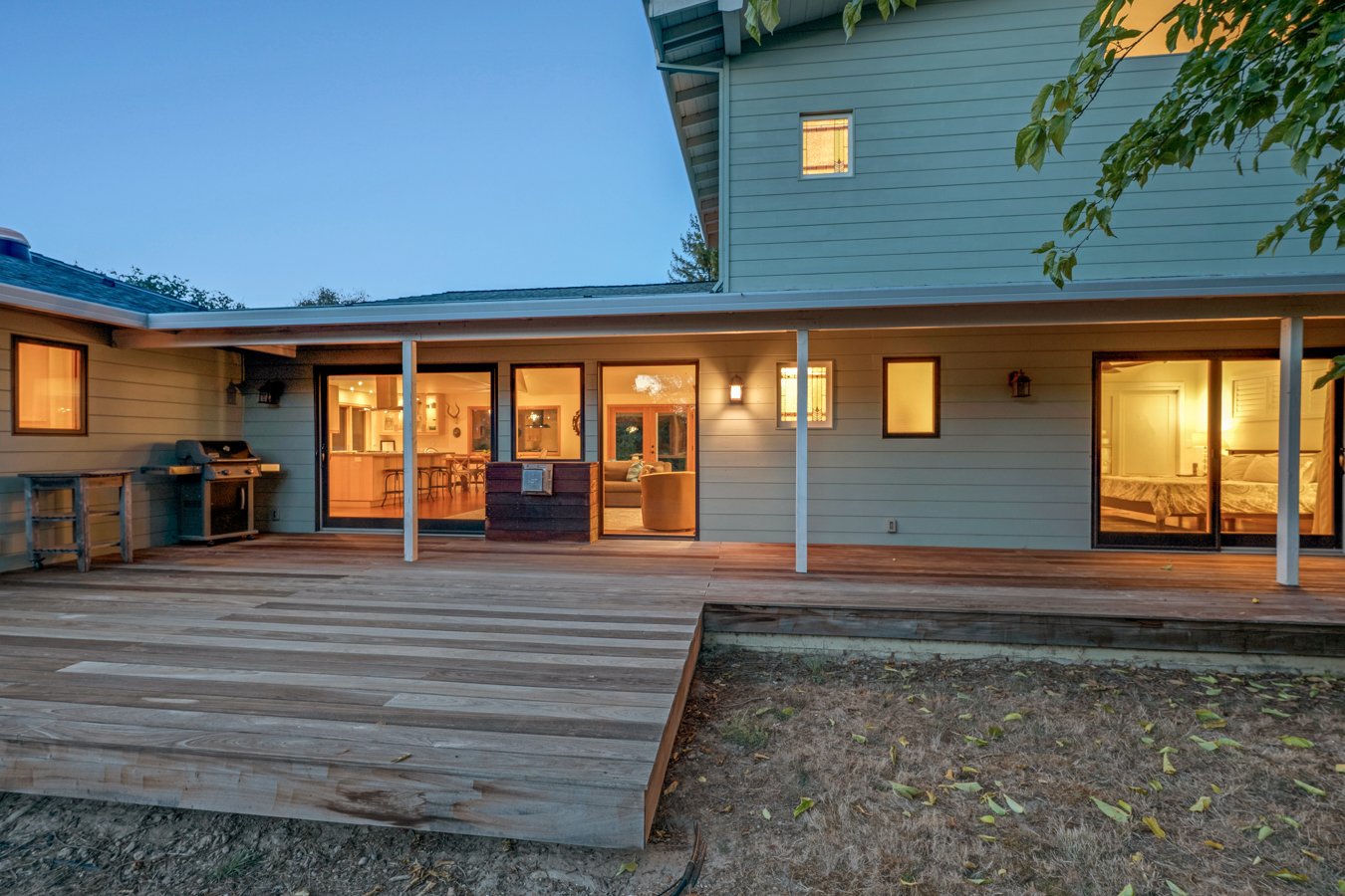 Residential house with a large wooden deck, sliding glass doors, and illuminated rooms inside, during dusk.