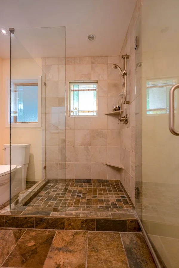 Bathroom shower area with beige tiled walls, small stained glass window, and a built-in corner shelf. Blue and brown tiled floor with a glass door, showing part of a toilet and a window with frosted glass.