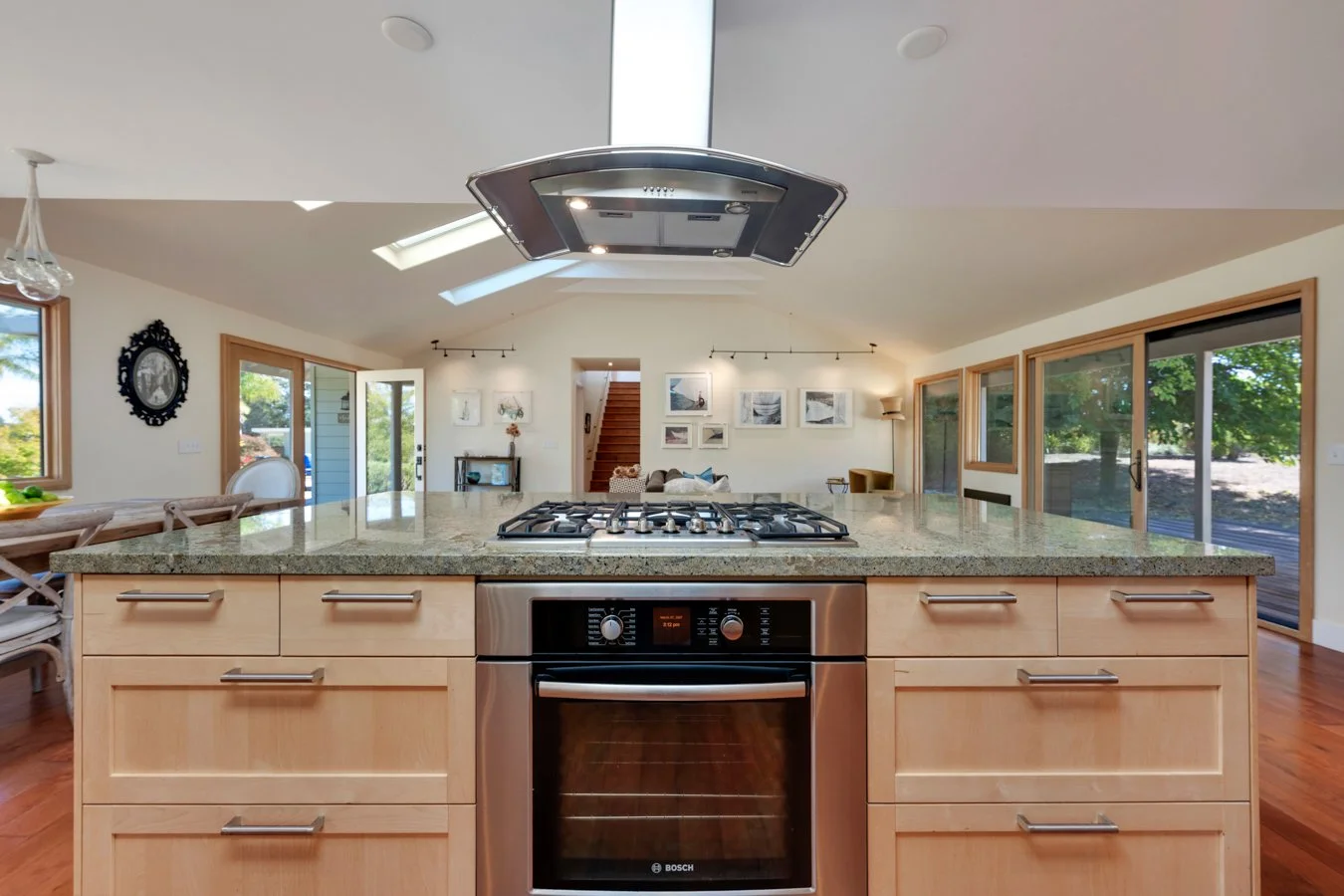 A modern kitchen with a central island, gas cooktop, built-in oven, granite countertops, wooden cabinets, large sliding glass doors, windows, and a ceiling with skylights.