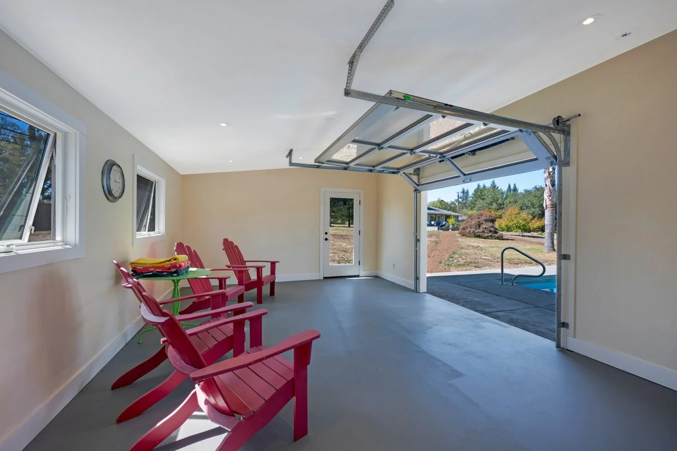 Bright garage with open door leading to backyard, three pink Adirondack chairs, small green table with folded towels, and windows showing trees outside.