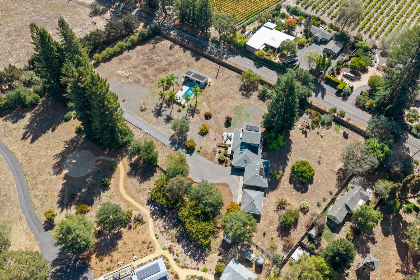 Aerial view of a residential backyard with a small pool, shaded seating area, garden, and trees, surrounded by neighboring houses and farmland.