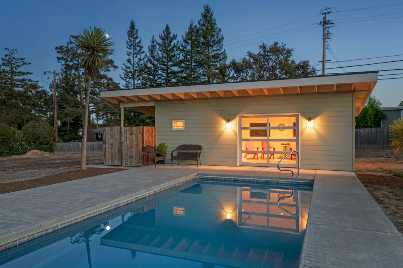 A backyard with a swimming pool, house with illuminated interior, outdoor seating, and tall trees in the background at dusk.