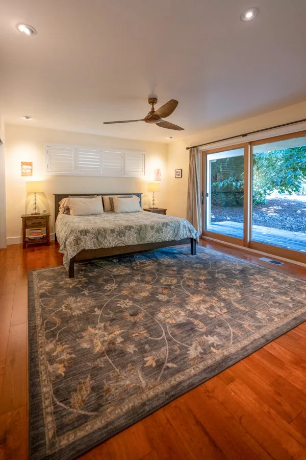 A cozy bedroom featuring a large bed with pillows and a floral bedspread, two nightstands with lamps, a large area rug with floral patterns, a ceiling fan, and sliding glass doors leading to an outdoor patio with greenery.