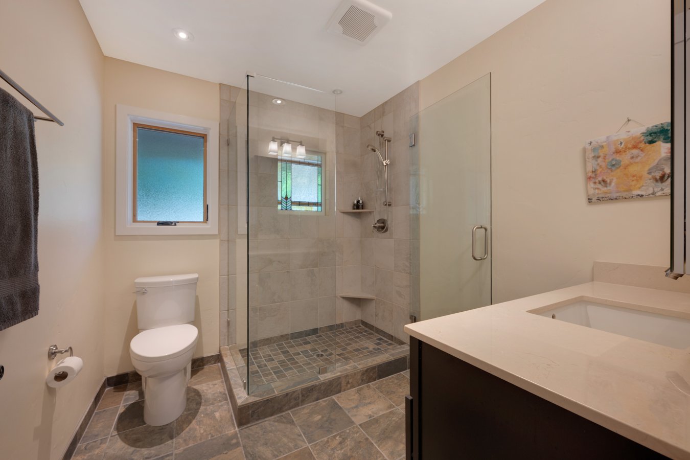 Bathroom with beige walls, frosted window, toilet, glass-enclosed shower with tiled interior, shower shelves, and a countertop with a sink.