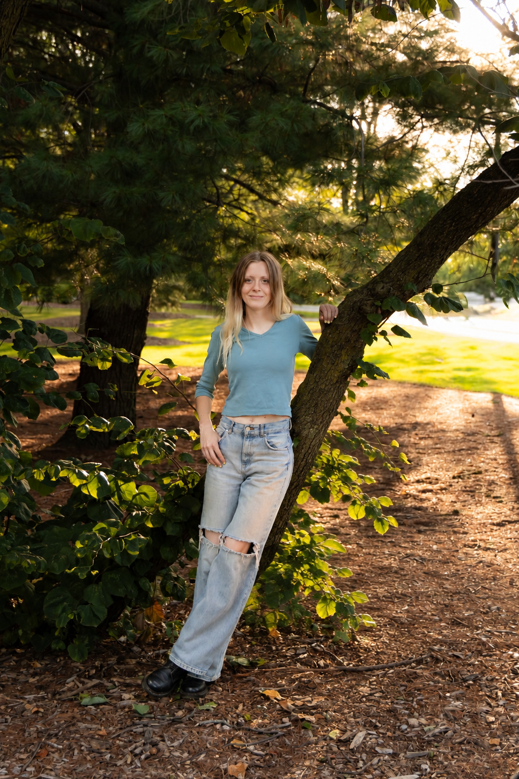 A young woman with blonde hair, wearing a blue crop top and ripped jeans, stands next to a tree in a park during sunset.