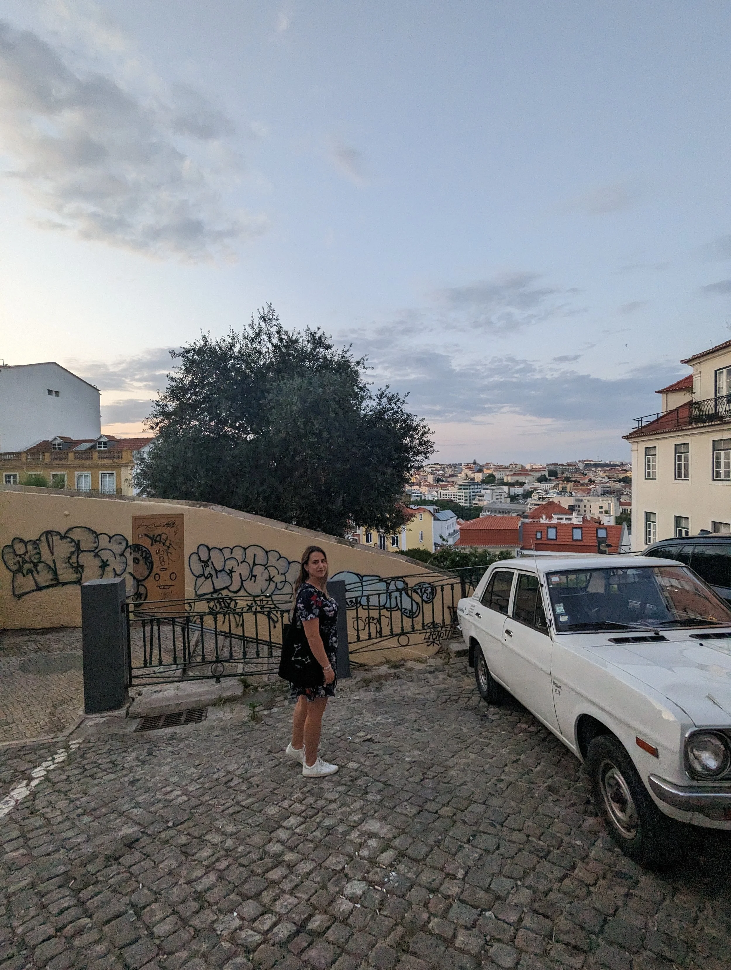 A woman standing on a cobblestone street at dusk, with a white car parked nearby, surrounded by urban buildings, trees, and graffiti on a wall.