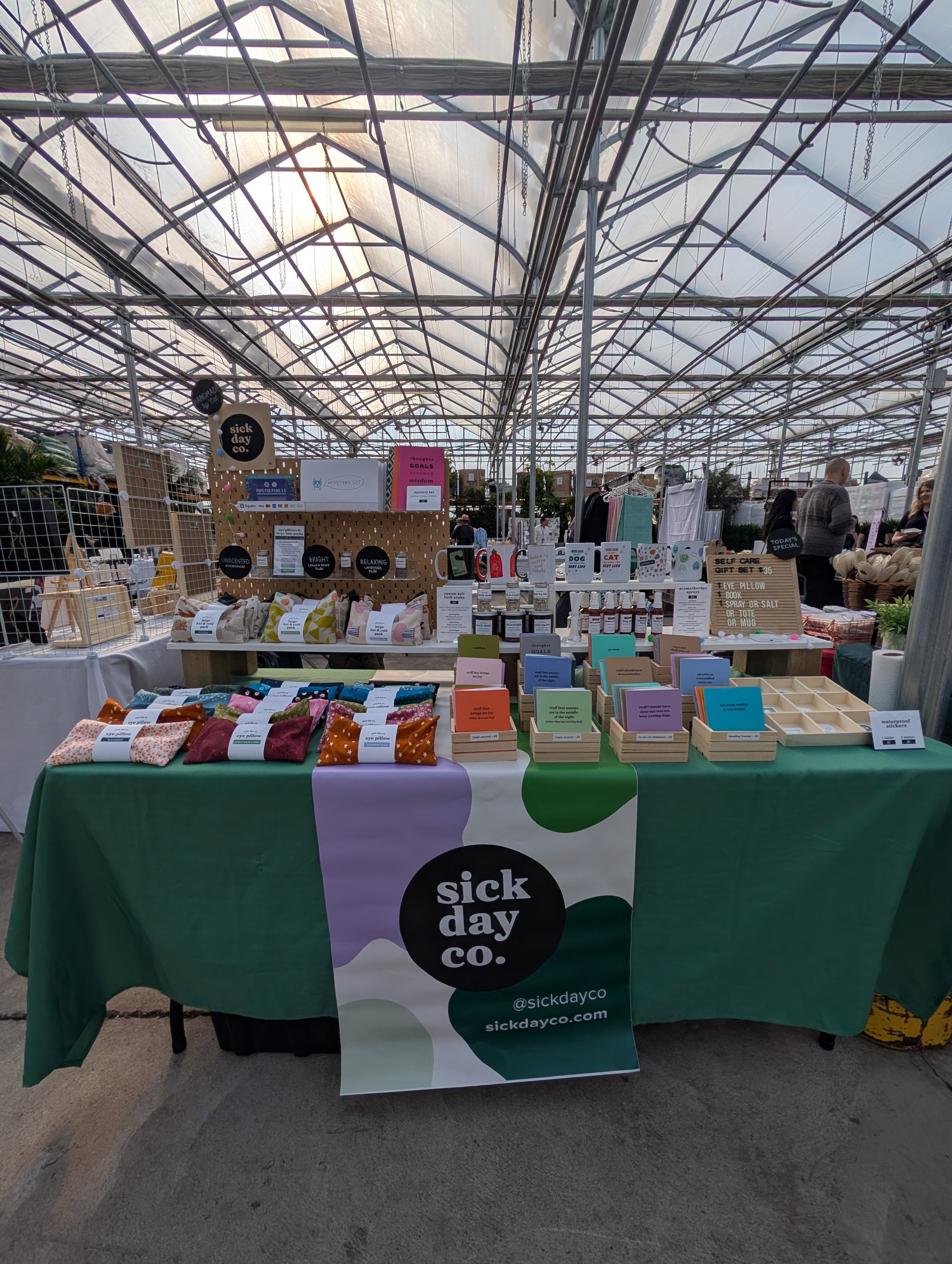 A booth display at a market with a green, white, and purple tablecloth bearing the logo 'sick day co.' and various wellness products. The background shows a greenhouse-style roof with metal framework.